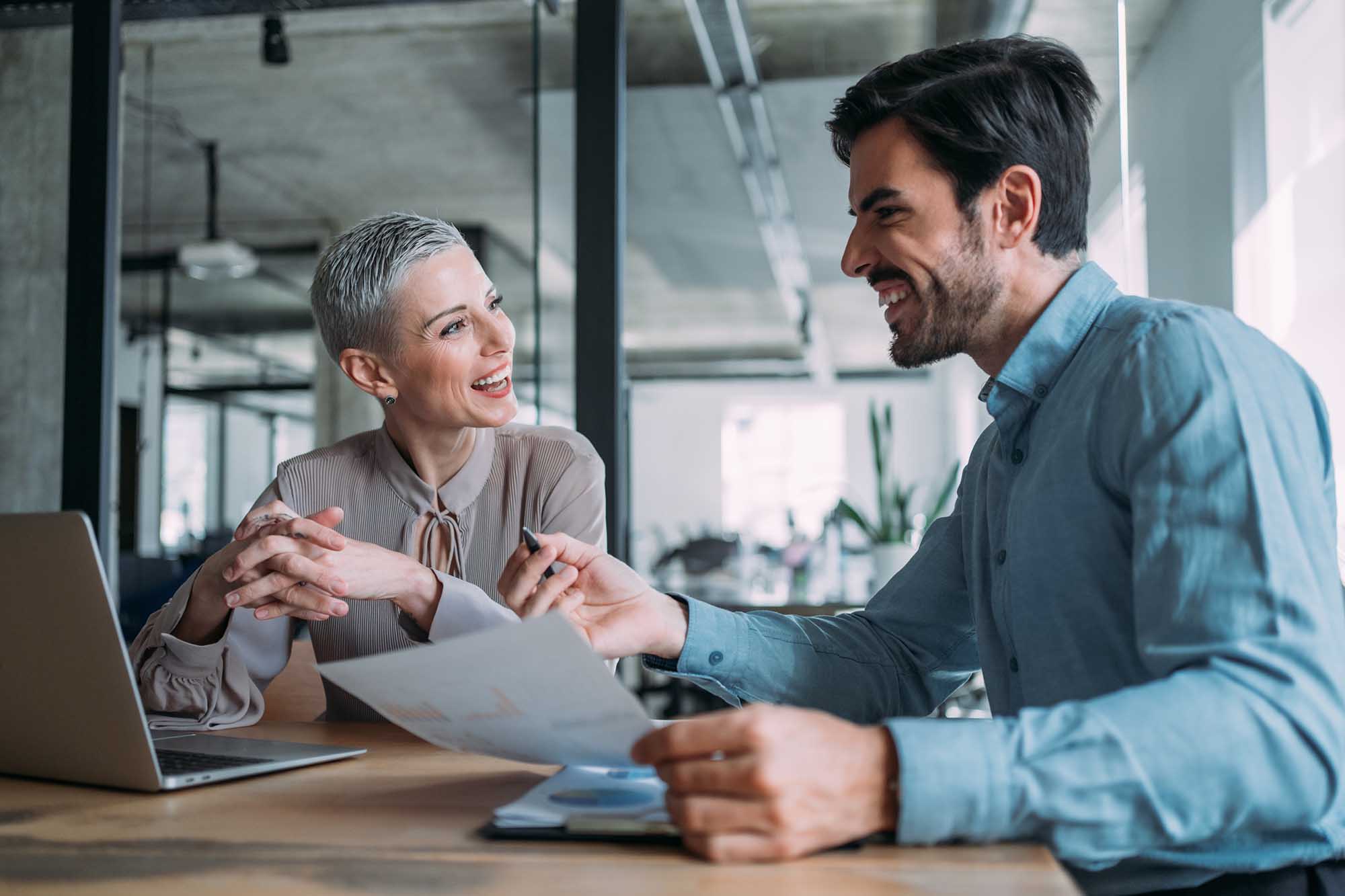 Shot of a two confident business persons sitting on a desk in the office and sharing ideas. Businessman and businesswoman in meeting using laptop and discussing business strategy. Business coworkers working together in the office.