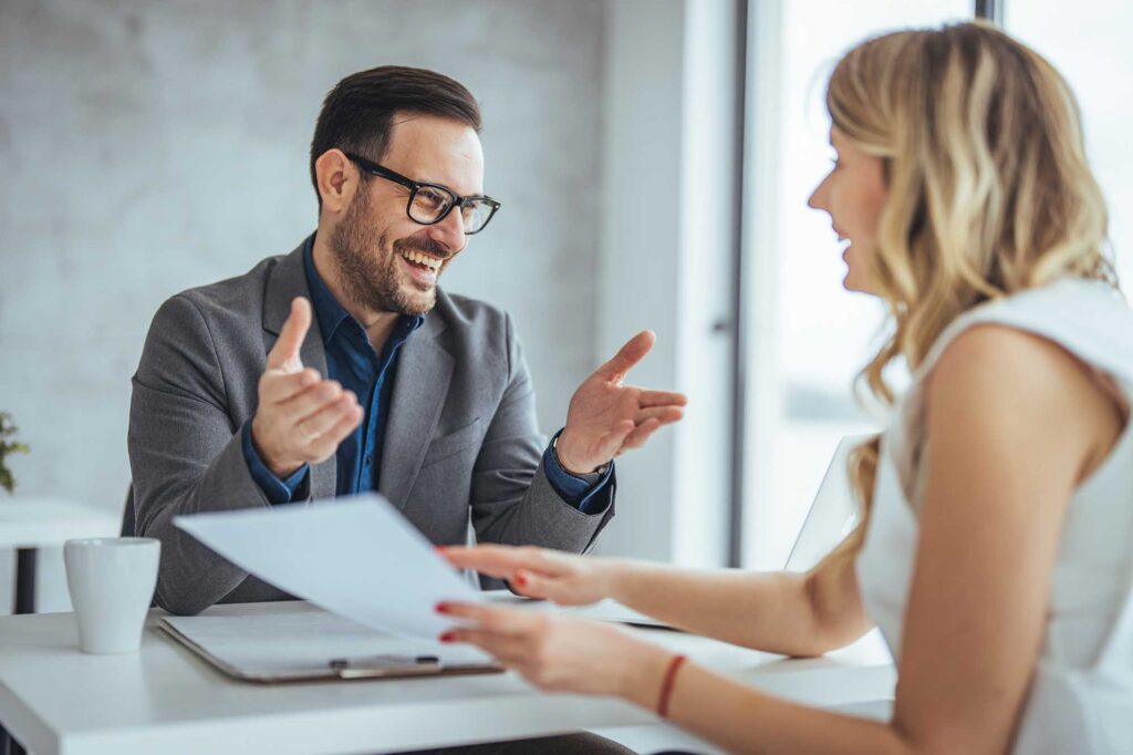A male and female in formal attire engage in a productive dialogue, sharing documents across the table in a well-lit office setting.