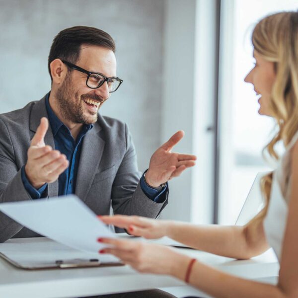 A male and female in formal attire engage in a productive dialogue, sharing documents across the table in a well-lit office setting.