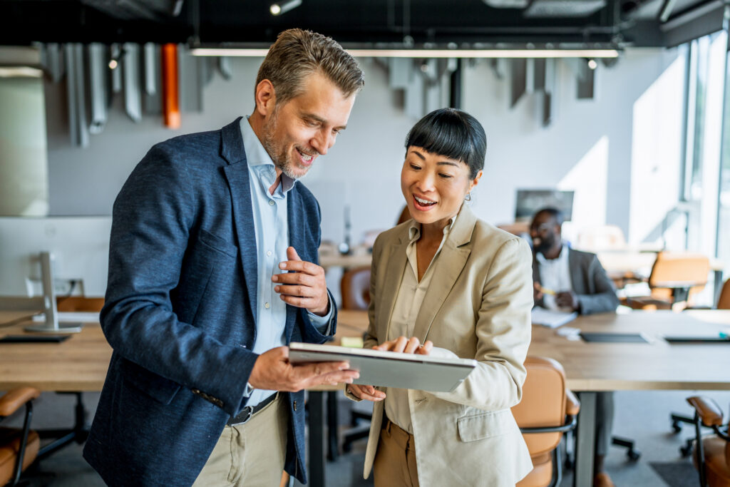 Two business colleagues are standing in a modern office, smiling and using a digital tablet together